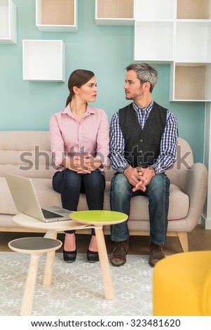Two coworkers are looking at each other, sitting on a sofa in a stylish vintage open space with pastels colors The grey hair man in checkered blue shirt and the woman in pink shirt are hands crossed