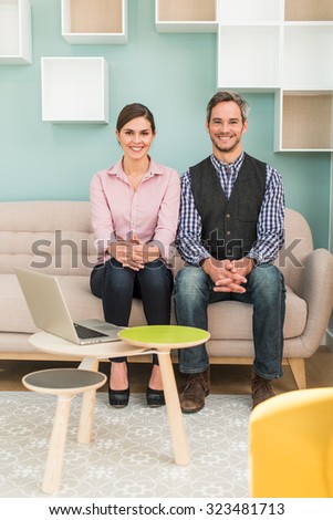 A grey hair man with checkered blue shirt and a woman in pink shirt are sitting on a sofa in a stylish vintage open space with pastels colors. They are smiling, looking at camera hands crossed.
