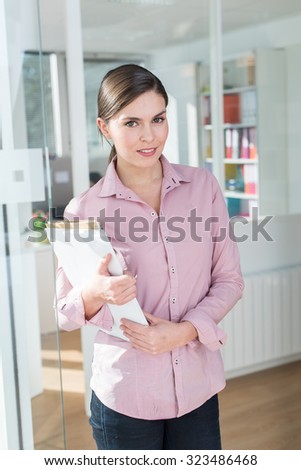 Portrait of a smiling thirty years old woman with her hair tied and wearing a pink shirt. She is holding folders and note books, standing in the hallway of a white office, next to a glass door.