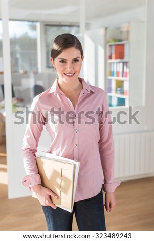 Portrait of a smiling woman standing in front of her office. She is wearing a pink shirt and jeans, holding folders in her hand. She is in a luminous open space with wooden floor.