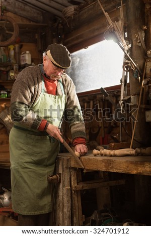 A grandfather is working on a wooden leg in his workshop. He is focused on his work, wearing a cap, a sweater and a green apron. The workshop is rustic with tools hanging from the ceiling