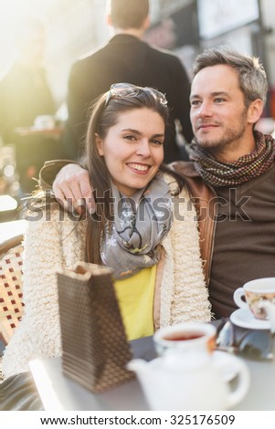 Trendy couple having fun and sitting at a terrace in the city center. The woman is wearing a woolen coat and a scarf The grey hair man has a beard and a leather coat Focus on the woman
