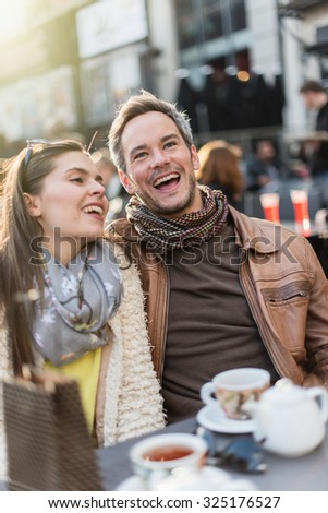 Trendy couple having fun and sitting at a terrace in the city center. The woman is wearing a woolen coat and a scarf The grey hair man has a beard and a leather coat A shopping bag is on the bar table