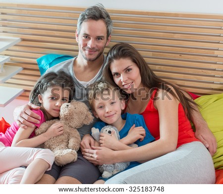 Parents and their two children are sitting in bed in the morning. The little girl and her brother are holding their cuddly toys. There is a wooden headboard behind them. They are looking at camera
