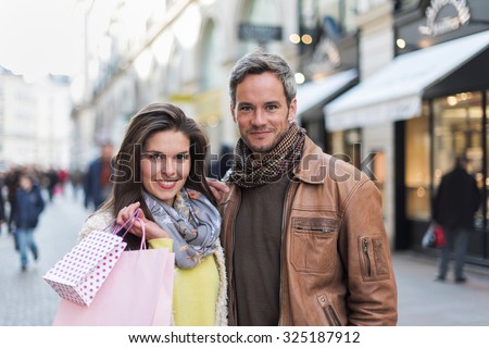 A trendy couple is standing arm in arm in the city center. They are in a cobbled car-free street. The woman is wearing a yellow shirt and pink shopping bags and the grey hair man has a leather coat