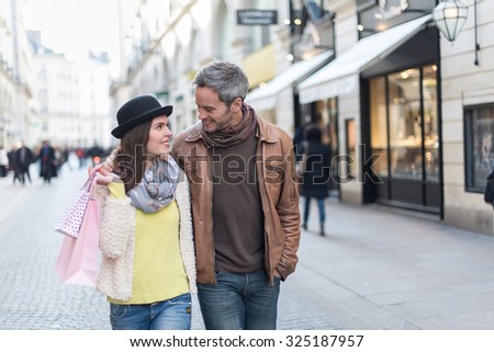 A trendy couple is walking arm in arm in the city center. They are in a cobbled car-free street. The woman is wearing a black hat and pink shopping bags and the grey hair man has a leather coat