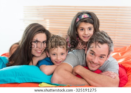 Portrait of a happy family having fun in bed They are looking at camera, laying one on top of another They are wearing casual clothes on a bright color bed. The little girl has a head band in her hair