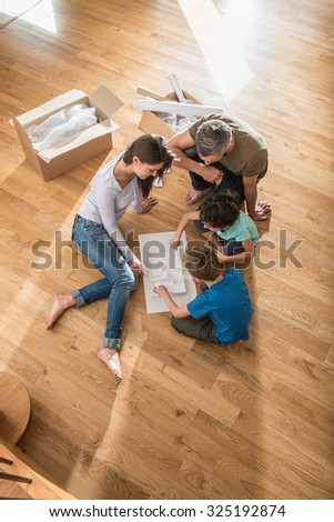 A mother and father are sitting barefoot on the floor of their new house. Their daughter and son are playing with the house model. They are surrounded by open cardboard boxes and palettes.