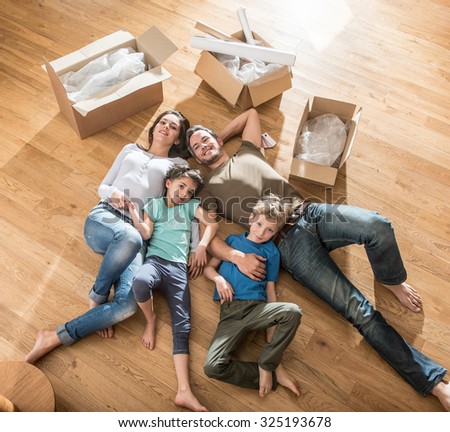 Top view of a happy family in casual clothes laying on the wooden floor of their new flat with cardboard boxes around them They are looking at camera The smiling parents are holding their two children