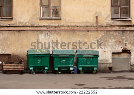 Three street green garbage can with wheels and folding lids. Near ragged wall.