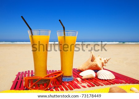 two orange juices, seashells and sunglasses on a deserted beach in a summer day with deep blue sky