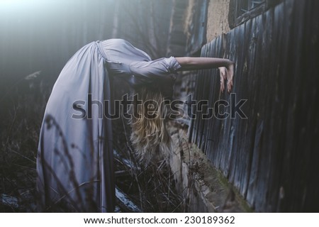 Blonde young model in a lavender maxi dress bends back in a creepy unnatural way in the dark forest