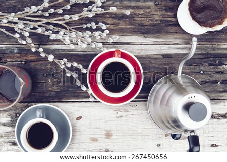 Two fresh morning cups of pour over coffee with saucers, a kettle, a dripper filled with filter and used coffee and a pussy willow on the old wooden table background
