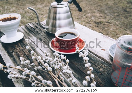 Fresh morning coffee in a red cup on the old wooden table with a kettle, coffee dripper and some pussy willow outdoor picnic