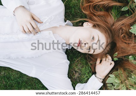 Beautiful redhead woman lying on the fresh green grass wearing a white vintage shirt