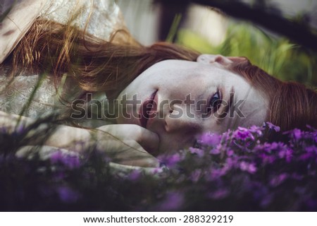 Beautiful sleepy redhead woman lying on the green grass among little purple flowers