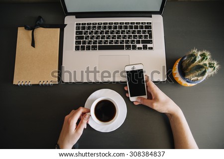 Top view of a business person holding a cup of fresh coffee and a smartphone, sitting at the desk. On the office table: laptop, cacti,sunglasses,smartphone, coffee and a paper notebook