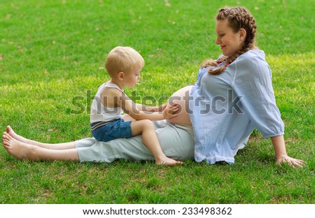 Pregnant woman and her son 2 yo have a rest on the green grass. Concept of motherhood and femininity