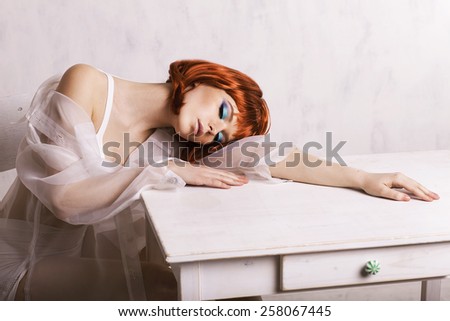 Young woman with red hair sitting near the table and thinking. Bob hair cut. Red hair. White table. Sitting and posing. Fashion model. Closed eyes.