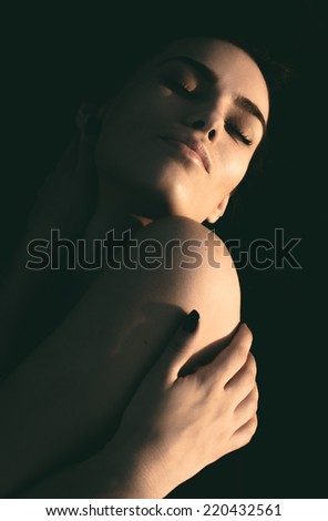 Artistic evocative dark portrait of a young woman with her hand wrapped around her body tilting her head back into the light with her eyes closed and a dreamy sensual expression