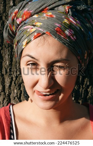 Close up Attractive Young Woman Winking with Tongue Out Leaning on Tree, Wearing Floral Printed Head Scarf.