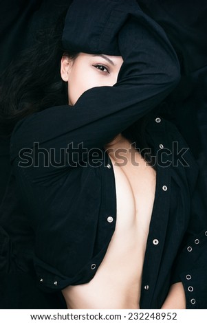 Close up Sexy Young Woman in Black Unbuttoned Long Sleeve Shirt, Showing her Cleavage, with One Arm Crossing her Face. Capture her While Lying on the Floor with Black Cloth Background.