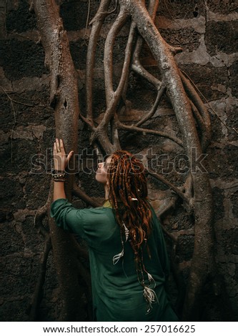 Rear View of Stylish Young Woman in Green Shirt with Dreadlocks Touching Tree Roots on the Wall.