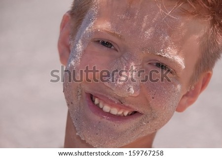 Teenager caucasian boy is smiling at the beach. His face is in the sand. Happy child is in summer day. Male model outside. Summer vacation concept. Close up.