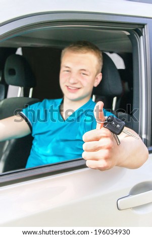 Car driver. Caucasian teen boy showing thumbs up and car key in the car window. Happy smiling young man behind the wheel. Travel and rental concept. Close up, outdoor.