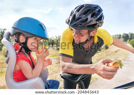 Happy father and son is eating lunch (snack) during bicycle ride. Child (boy) and man have biking helmets. The son is in the bicycle chair (seat). Caucasian smiling male models. Travel concept.