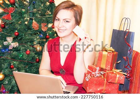 Young attractive girl is buying online. Happy woman with laptop, credit card and gift boxes on christmas tree background. Caucasian beautiful adult female in her 20s. New year concept.