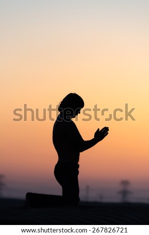 Praying man silhouette staying on his knees alone in desert at sunset