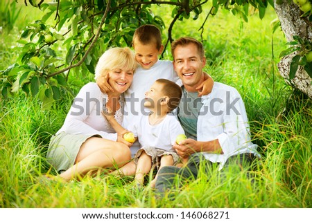 Happy Big Family Outdoors Having Fun. Family with two Children relaxing and Enjoying nature. Outside. Smiling Parents and Kids lying on green grass in the Orchard, Eating Apples and Laughing