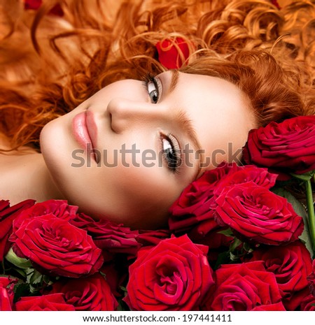 Beauty model girl with long curly red hair and beautiful red roses bouquet. Hairstyle with flowers. Fashion woman with Wavy healthy hair lying on beautiful roses. Permed hair