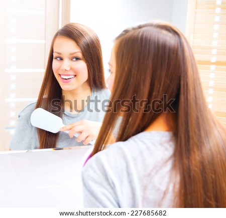 Beautiful smiling teenage girl is combing her long silky hair after getting up in the morning. Young attractive happy woman is brushing her brown hair enjoying them in the mirror and admiring herself