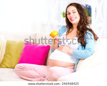 Pregnant Happy Woman sitting on a sofa and eating apple. Mom Expecting Baby. Healthy eating, diet, dieting. Pregnant Woman Belly. Pregnancy. Baby Shower