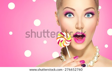 Beauty fashion model girl Eating colourful lollipop. Surprised Young funny woman with pink nail art and makeup over pink polka dots background