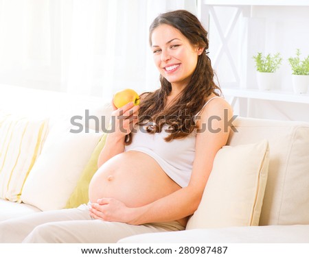 Pregnant Happy Woman sitting on a sofa and eating apple. Mom Expecting Baby. Healthy eating, diet, dieting. Pregnant Woman Belly. Pregnancy. Baby Shower