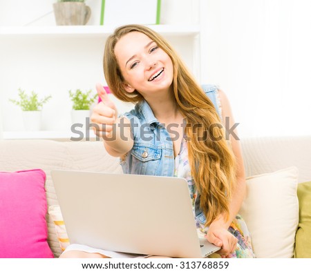Pretty teenage girl sitting on sofa at home with her laptop, making thumb up gesture. Beauty girl enjoying the networks by laptop computer, laughing student.