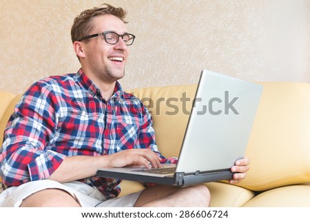 Handsome hipster guy looking at his laptop computer in his hans and smiling - Man in glasses wearing red shirt laughing against computer about a funny moments picture on the sofa - chilling concept