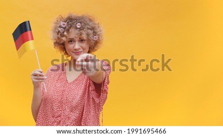 Attractive young girl holding a small German flag in her hand. Pointing finger at the camera. Isolated over yellow background. Studio shot. Concept of Learning German. Foto stock © 