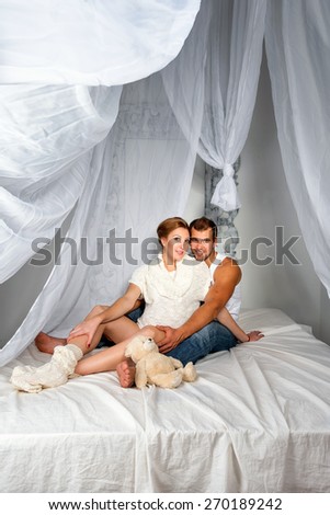 Happy Pregnant Couple dressed in white embrace each other sitting on a bed with a canopy on a white background