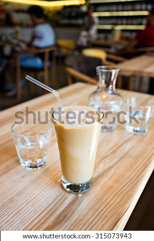 A cup of cold ice coffee with milk and tube to drink in a glass on wooden background with carafe of water