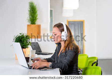 Young successful businesswoman in headphones with laptop computer surfing the internet at modern bright office. Woman using tablet computer and listening to the music during coffee break