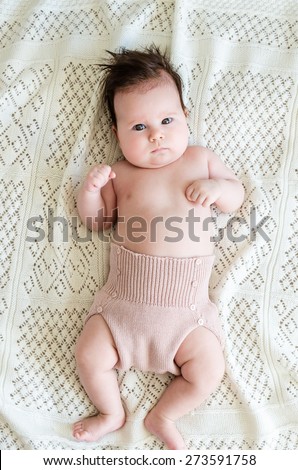 Cute sweet newborn baby girl in woolen shorts with big beautiful hazel brown eyes and curious glance lying on white woolen blanket on a bed at home, top view.