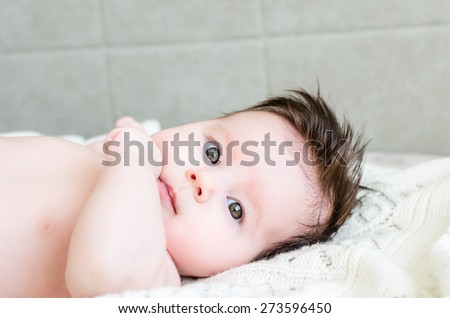 Cute sweet little newborn baby girl lying on white woolen blanket on a bed at home and looking with big beautiful hazel brown eyes and curious glance