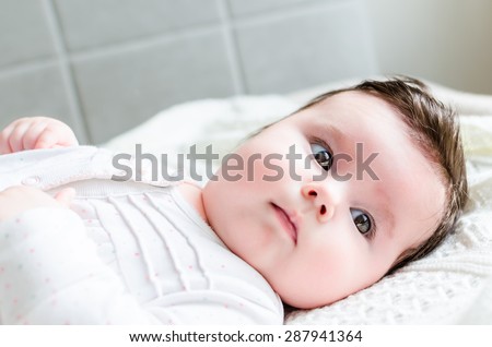 Portrait of cute sweet little newborn baby girl with black brunette hair in nice white spotted romper suit looking with big beautiful hazel brown eyes, lying on white woolen blanket on a bed at home