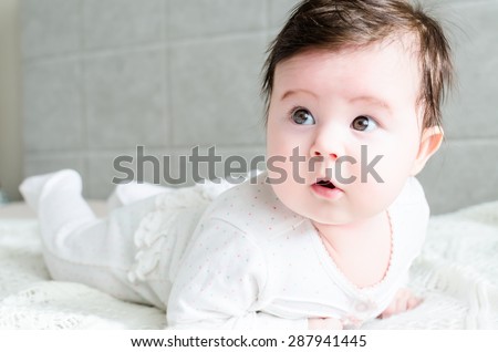 Cute sweet little newborn baby girl with black hair in nice white spotted romper suit looking with big beautiful hazel brown eyes, with opened mouth and lying on white woolen blanket on a bed at home