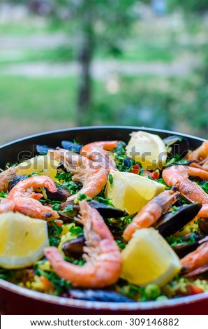 Close up fresh juicy prepared classic spanish dish paella with seafood, blue shell mussels, shrimps, rice with saffron spice, vegetables, tomatoes, smoked sausages and lemon with parsley in frying pan