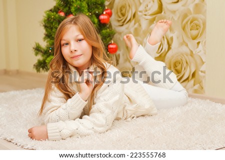 Girl lying on the carpet, looking at the camera and smiling. A child in a warm white sweater. Christmas tree. Teen in New Year clothes.